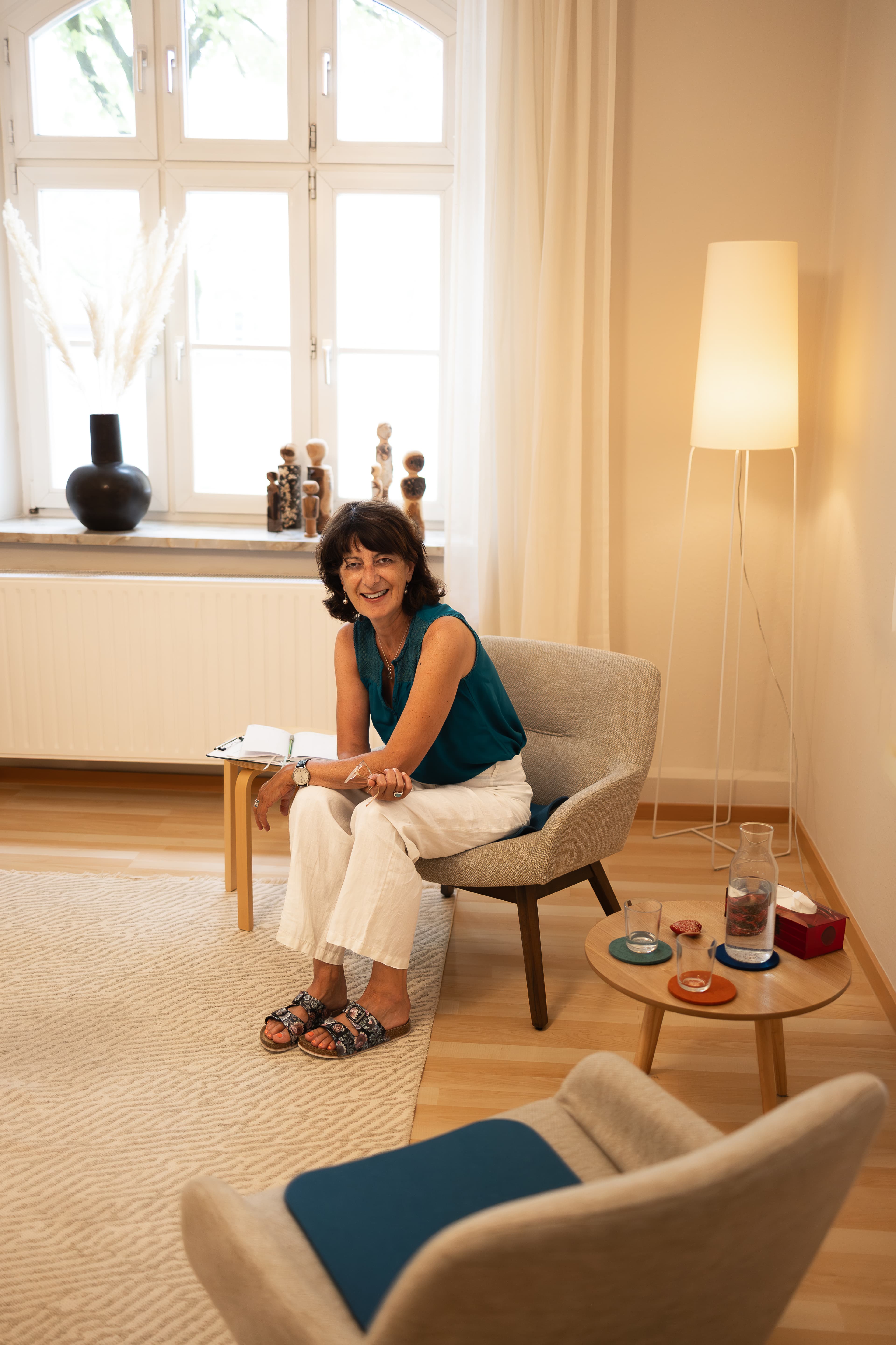 Corinne Charvet sitzt lächelnd im Sessel ihres Therapieraums in München-Neuhausen, daneben ein Beistelltisch mit aufgeschlagenem Notizbuch